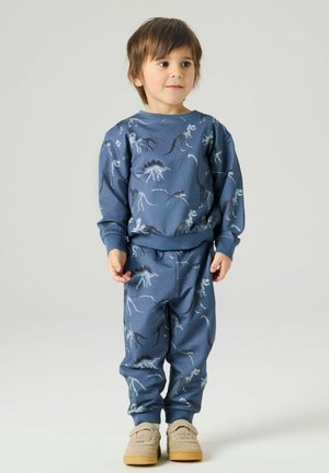 Noor poiss, kellel on seljas sinise dinosauruse mustriga dressipluus ja sobivad püksid, seisab ühevärvilise heleda tausta ees.