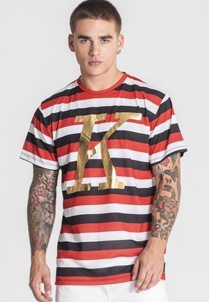 Giovane uomo con capelli corti che indossa una maglietta a righe rossa, nera e bianca con una grande lettera "K" dorata sul davanti e braccia tatuate.