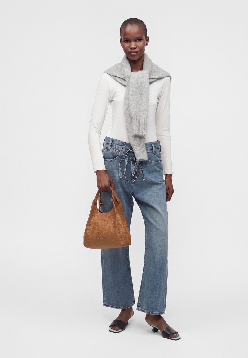 Femme portant un haut blanc à manches longues, une écharpe grise, un jean bleu ample, des talons noirs ouverts à l'avant, tenant un sac à main en cuir marron sur un fond uni.