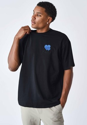 T-shirt en coton noir avec un graphisme de cœur bleu sur la poitrine et un texte brodé. Manches courtes avec une coupe décontractée.