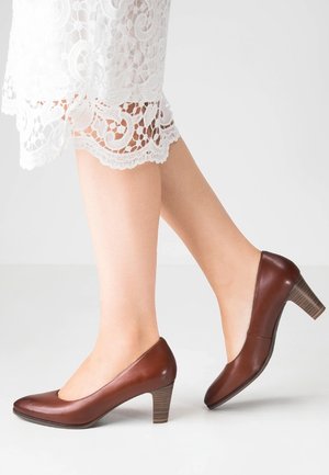 Nogi w brązowych skórzanych butach na średnim obcasie i w białej koronkowej sukience, stojące na jasnoszarym podłożu na tle jednolitego, białego tła.