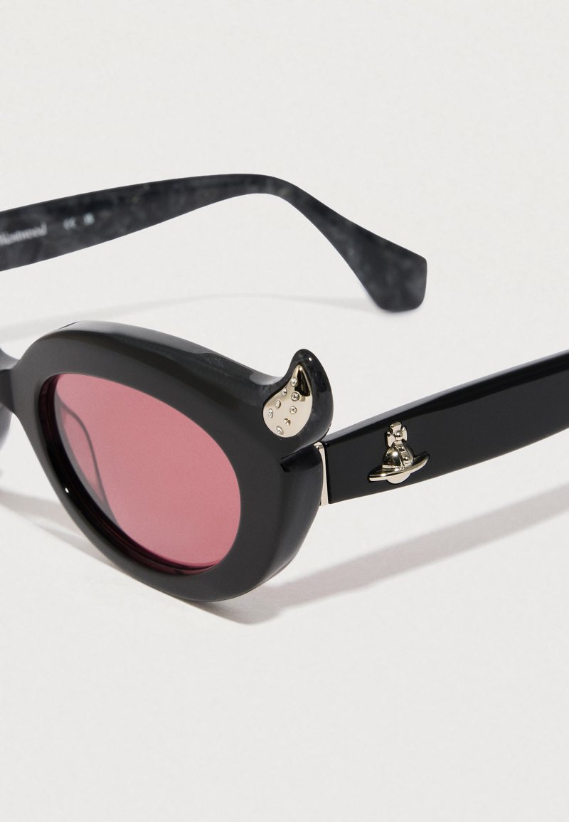 Black sunglasses with round pink lenses, featuring a curved horn-like detail on the frame and metallic accents near the temples.