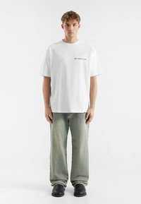 White cotton t-shirt with a crew neckline and a black "DON'T WASTE CULTURE" print on the left side, paired with light green jeans and black shoes.