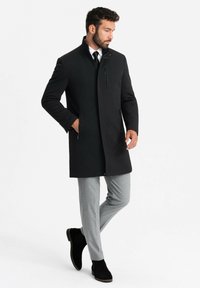 Cappotto da uomo in lana nera con colletto alto, tasca con zip frontale e tasche laterali, abbinato a pantaloni grigi chiari e stivali neri alla caviglia.