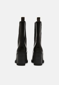 Une paire de bottines en cuir noir à talons blocs, avec des panneaux latéraux élastiques et des languettes, présentée de dos sur un fond blanc.