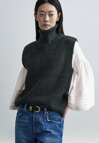 Donkergroene geribbelde coltrui gecombineerd met een lichtroze blouse met geplooide mouwen, gedragen met blauwe spijkerbroek en een zwarte riem.