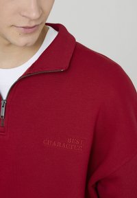 Pull-over rouge avec un col côtelé, une fermeture éclair et le texte brodé « BEST CHARACTER » sur la poitrine. Confectionné en tissu texturé.