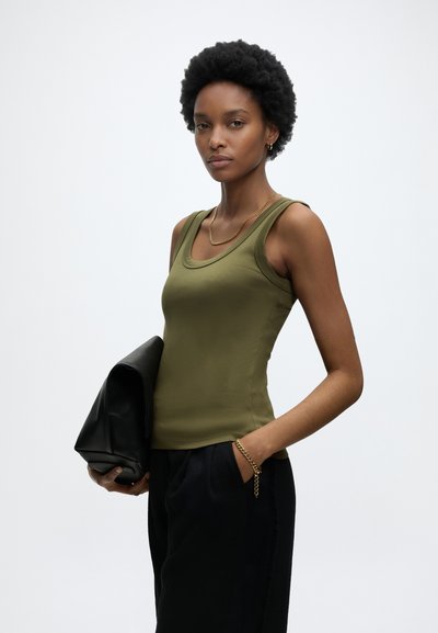 Νεαρή γυναίκα με κοντά σγουρά μαλλιά, φοράει χακί αμάνικη μπλούζα και μαύρο παντελόνι, κρατάει μαύρο δερμάτινο τσαντάκι και φοράει χρυσά κοσμήματα.