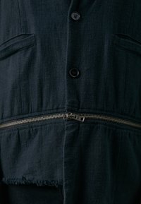 Dunkelblaue Stoff-Jumpsuit mit einem Reißverschluss vorne, zwei Brusttaschen und einem ausgefransten Saumdetail mit mattschwarzen Knöpfen.
