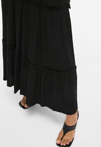 Jupe maxi noire à volants avec un tissu texturé et un ourlet festonné, associée à des sandales noires à une bride présentant une semelle plate.