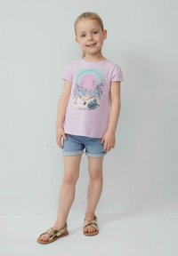 Hellblaues kurzes T-Shirt mit grafischem Design, das Palmen und eine Hängematte zeigt, kombiniert mit Jeansshorts und goldenen Sandalen.