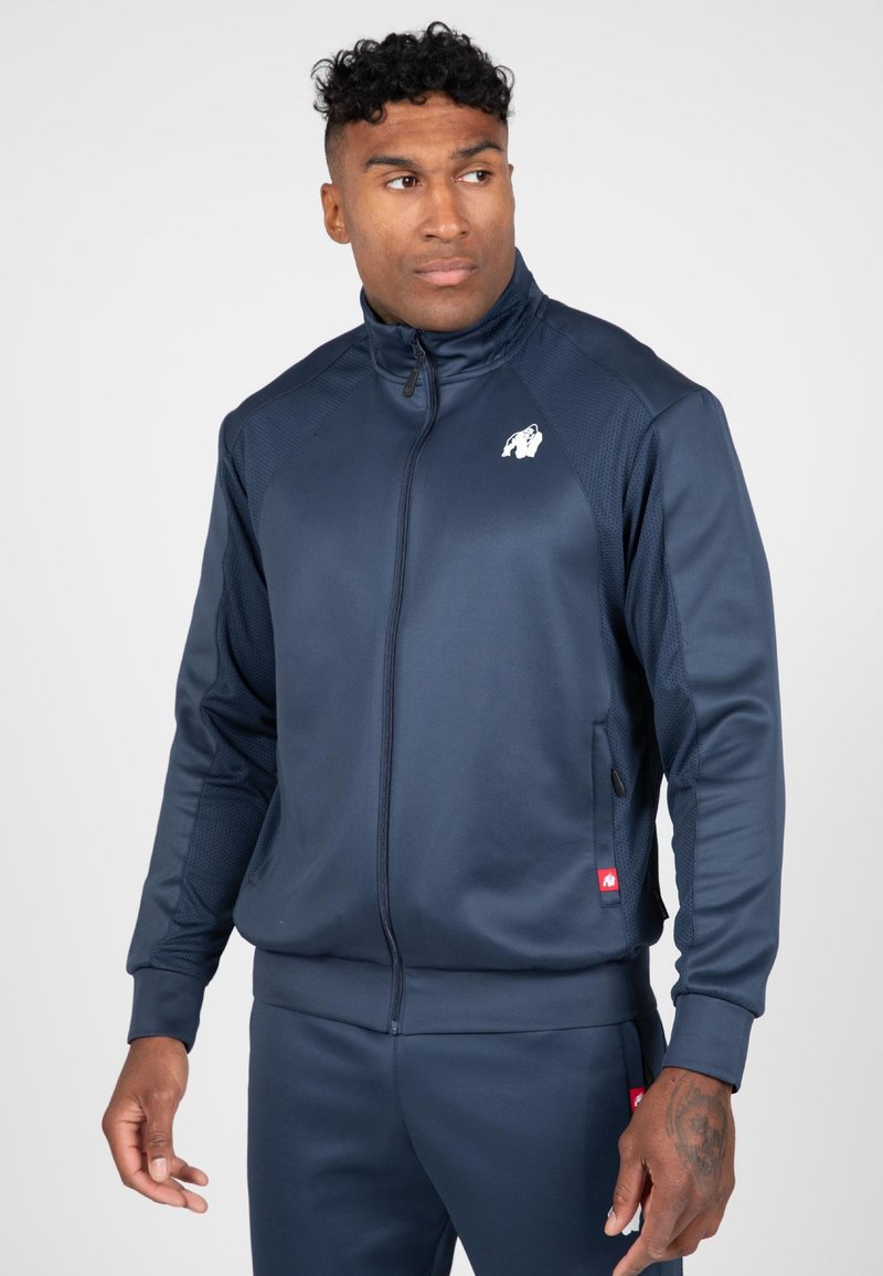 Navy zip-up jack met gladde stof, met textuurpanelen aan de zijkanten, een logo op de borst en ribbelboorden. Bijpassende broek zichtbaar.