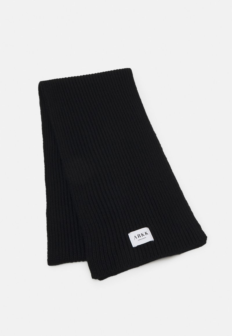 Svart ribbestrikket skjerf laget av stoff med en teksturert overflate, med en hvit etikett som har "ARKK"-merking. Lang og rektangulær form.