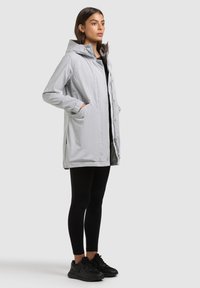 Femme portant un manteau à capuche gris clair sur des leggings noirs et des baskets noires, debout les mains dans les poches du manteau, regardant droit devant.