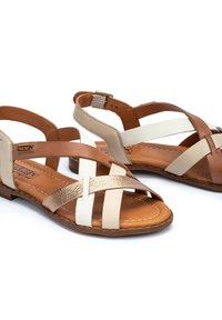 Bruine en beige leren sandalen met een kruislings ontwerp, voorzien van een metallic gouden accent en een vlakke zool. Verstelbare enkelband.