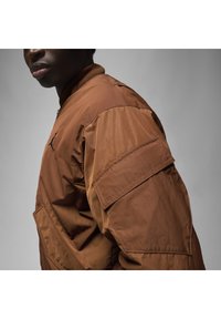 Giacca bomber marrone con una texture liscia, dotata di colletto a coste, chiusura con zip e tasca con patta sulla manica.
