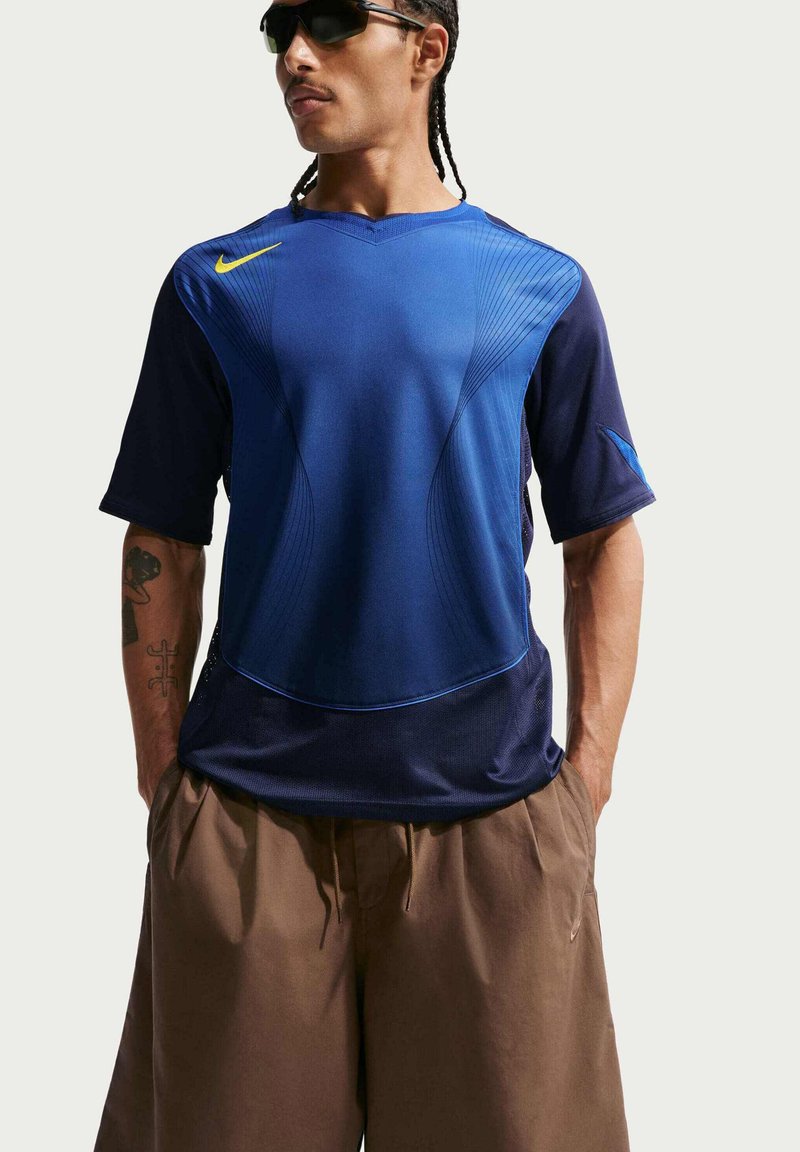 Hombre con gafas de sol, camiseta deportiva azul de Nike con logo amarillo, pantalones sueltos marrones, de pie con las manos en los bolsillos contra un fondo simple.