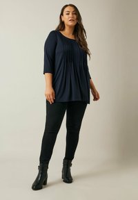 Marine tuniek met plooien, ronde halslijn en driekwart mouwen. Draagt het met zwarte skinny jeans en zwarte haklaarzen.