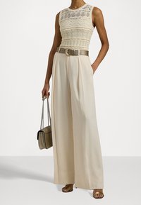 Kvinna iklädd krämfärgade vidbyxor och en ärmlös krämfärgad virkade topp, hållandes en metallisk handväska med kedjerem.