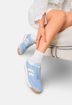 Personne assise sur un banc rembourré portant des baskets bleu clair avec des rayures crème, des chaussettes blanches et une chemise blanche à manches longues.