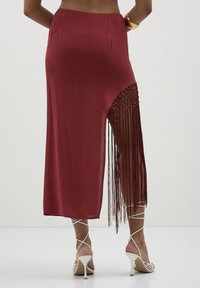 Femme portant une jupe midi bordeaux avec une fente à franges nouées sur le côté, des chaussures à lacets blanches, et un bracelet en or.