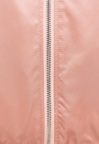 Jasnoróżowy materiał kurtki o gładkiej teksturze, z centralnym metalowym zamkiem błyskawicznym i kontrastującym szyciem wzdłuż szwu.