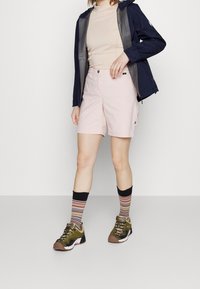 Světle růžové šortky s uvolněným střihem, spojené s béžovým topem a tmavě modrou bundou. Obsahují víceduhové pruhované ponožky a olivově zelené boty.