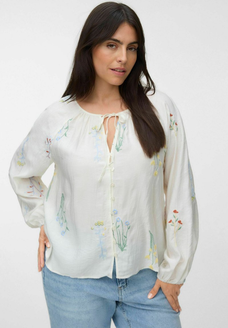 Femme aux longs cheveux foncés portant un chemisier blanc boutonné avec broderies florales et un jean bleu clair sur fond blanc.