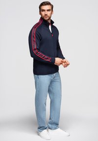 Marineblaue Zip-Jacke mit hohem Kragen; rote Streifen entlang der Ärmel; kombiniert mit hellblauen Jeans und weißen Sneakers, was einen sportlichen Look ergibt.