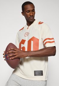 Kansas City Chiefs -jalkapallo paita vaaleassa värissä, jossa on oransseja yksityiskohtia, ja rohkea numero 60. Paitaa pitävä henkilö pitää käsissään jalkapalloa, jossa näkyvät pinnat ja logot.
