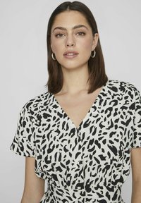 Sukienka z krótkim rękawem i dekoltem w serek w białym materiale z czarnym wzorem zwierzęcym. Zawiera guziki na przodzie dla dodatkowego akcentu.