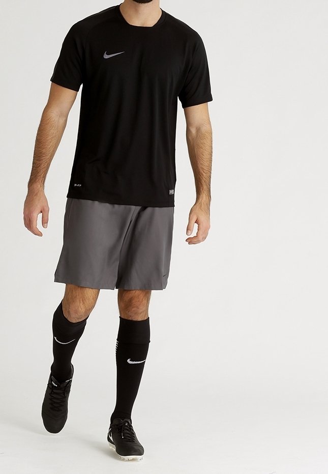 Homme marchant portant un t-shirt de sport Nike noir, un short gris, des chaussettes hautes noires et des crampons noirs sur un fond blanc uni.