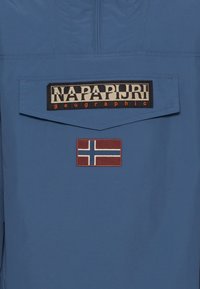 Blue jacket featuring a large black label with "NAPA PIVIRI" text and a brown patch with the Norwegian flag. Smooth fabric texture.