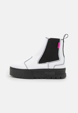 Bottine blanche en cuir avec panneau latéral élastique noir, semelle épaisse noire, coutures blanches et petite étiquette rose sur fond uni.