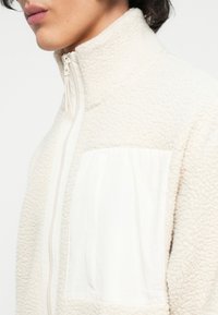 Veste en polaire écrue avec un col haut, fermeture éclair et une poche en tissu blanc contrastant. Surface texturée au toucher doux.