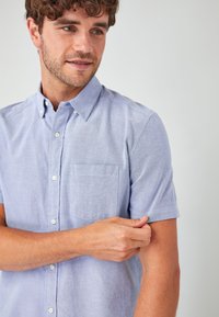 Camisa de botones de manga corta, color azul claro, en una tela suave, con un bolsillo en el pecho y un diseño de cuello clásico.