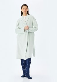 Mujer vestida con un albornoz verde claro sobre una camiseta blanca y pantalones de pijama azul marino con un patrón de ciervos, de pie descalza sobre un fondo blanco.