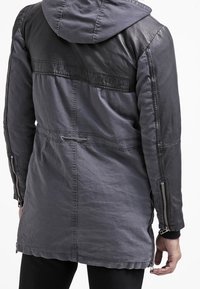 Grå jacka med en svart läderdetalj, huva och dragkedjor på sidofickorna. Har ett texturerat tyg och justerbar dragsko i midjan.