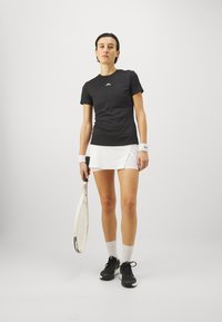 Juodas sportinis marškinėliai trumpomis rankovėmis su balta logo, derinami su balta teniso sijonu. Juodi sportbačiai ir apyrankės puikiai užbaigia įvaizdį.