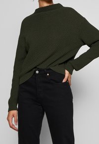 Sweter dziany w ciemnozielonym kolorze z wysokim kołnierzem, ściągaczami przy mankietach i teksturowanym wykończeniem, w zestawieniu z czarnymi spodniami z kieszenią z przodu.