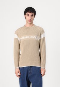 Beige gerippter Pullover mit weißem Streifen und Logo. Ausgestattet mit langen Ärmeln und einem runden Halsausschnitt, kombiniert mit lockeren blauen Denim-Hosen.