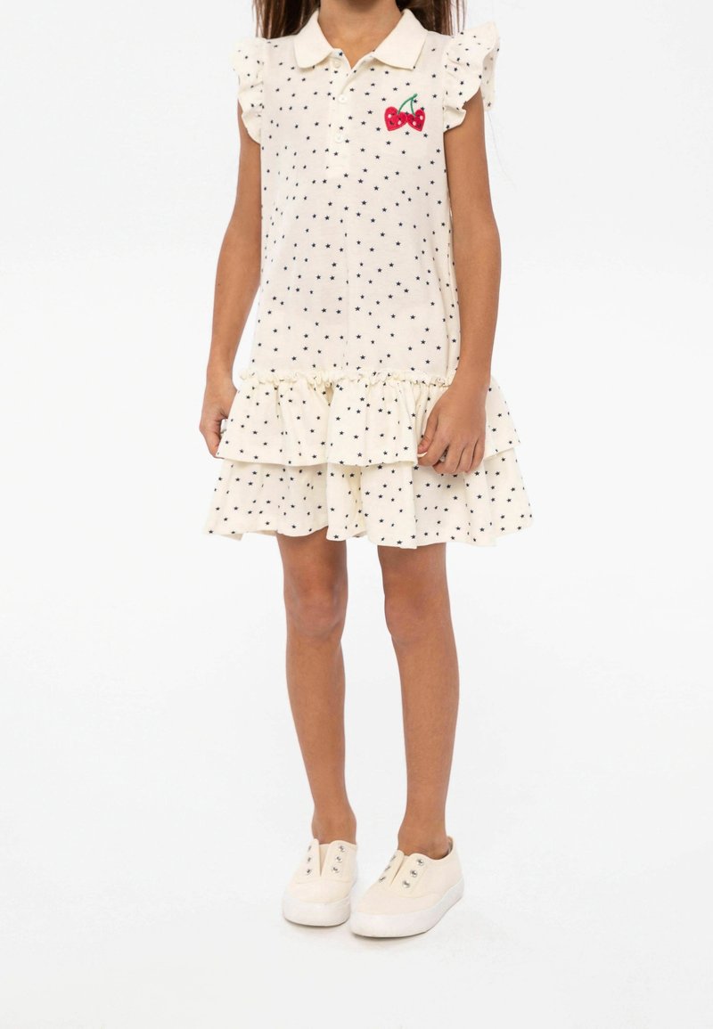 Fille portant une robe crème sans manches avec des motifs d'étoiles noires, une broderie de cerise sur la poitrine, une jupe à volants et des chaussures blanches à enfiler.