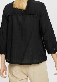 Blouse noire avec manches bouffantes trois-quarts, tissu texturé, bordure en dentelle festonnée le long de l'encolure et fentes latérales à l'ourlet.