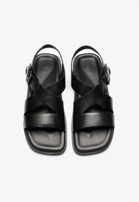 Sandales en cuir noir avec des brides croisées, une texture lisse, un dos ouvert, un bout carré et des détails de boucle ajustable sur le côté.