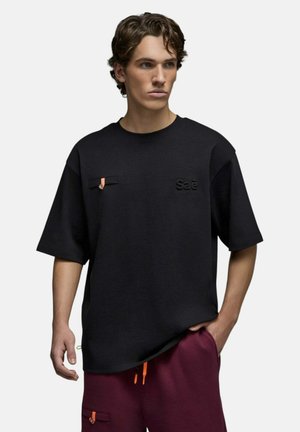 Jeune homme aux cheveux bouclés portant une chemise noire à manches courtes avec le logo "Sae" et un pantalon bordeaux avec des cordons orange, main dans la poche.