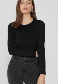 Schwarzes Langarmoberteil aus elastischem Material, mit Rundhalsausschnitt und tailliertem Design. Kombiniert mit hochgeschnittenen dunkelgrauen Jeans.