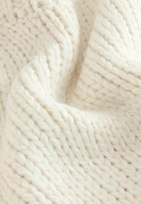 Gruby, off-white dzianinowy materiał o grubej strukturze włókna i miękkim, przytulnym wyglądzie. Wzór przedstawia przeplatające się szycia.