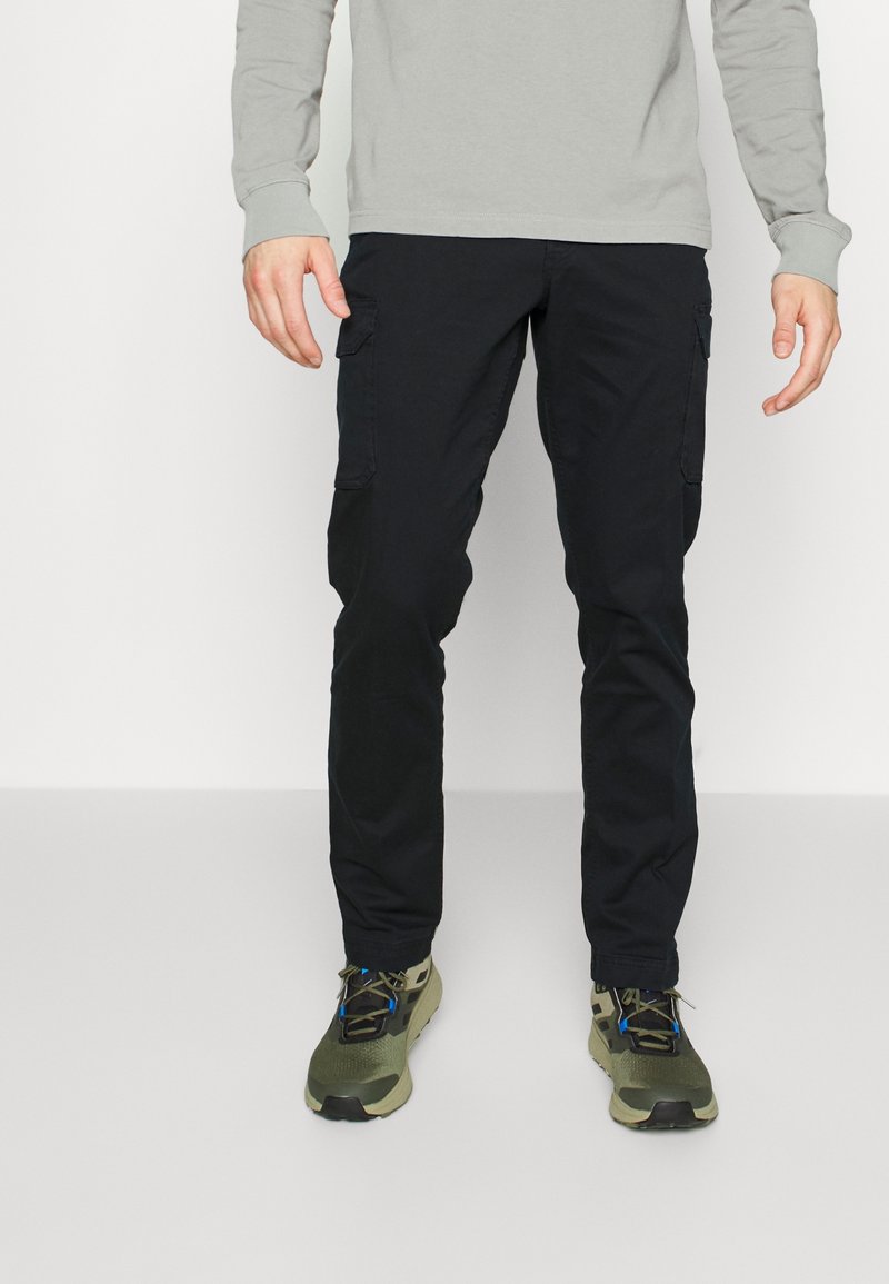 Pantalon cargo noir avec poches latérales, fabriqué en tissu robuste. Présente une coupe décontractée et des coutures visibles. Associé à des chaussures de sport vert olive.