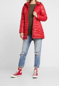 Chaqueta de plumas roja con cierre delantero, capucha y textura acolchada. Llevada sobre un suéter verde oscuro y combinada con unos jeans de mezclilla azul claro desgastados y zapatillas altas rojas.