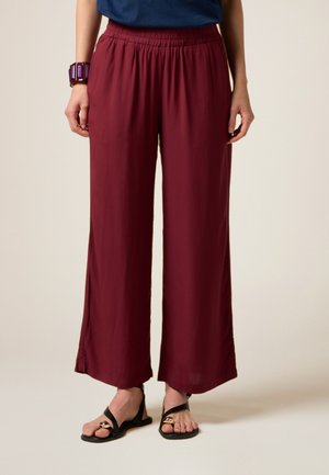 Pantaloni a gamba larga color bordeaux realizzati in tessuto leggero con una vita elasticizzata. Abbinati a sandali neri e a un braccialetto viola.